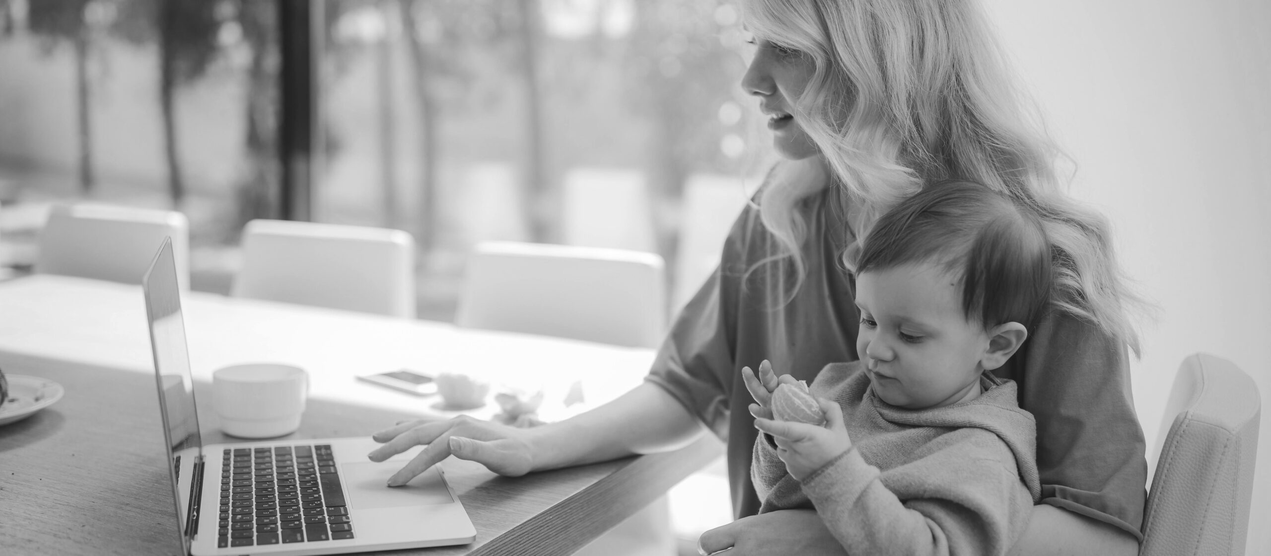 A woman working on a laptop while holding her toddler who is eating a mandarin